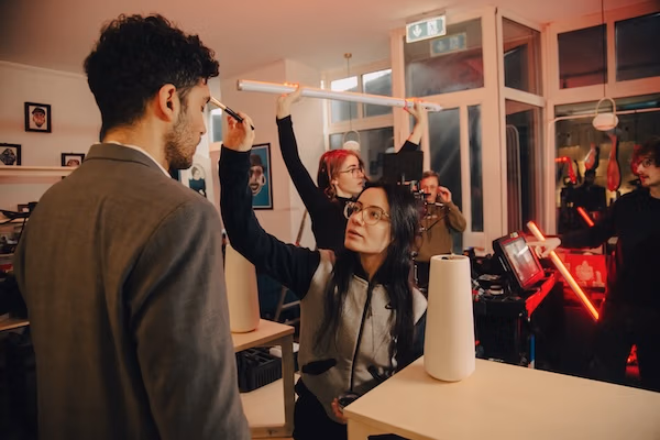 Behind-the-scenes-Szene am Filmset: Eine Maskenbildnerin schminkt einen Darsteller, während Crewmitglieder Licht und Kamera vorbereiten - für eine Spreadfilms Produktion