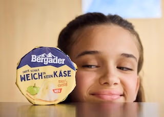 Kind blickt zufrieden auf eine Bergader-Weichkäse-Packung, die vor ihr auf dem Tisch steht- für das Bergader Werbevideo von Spreadfilms