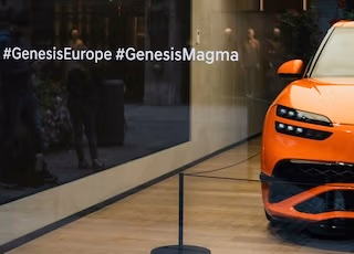 Ein orangefarbenes Auto steht in einem modernen Showroom , die Hashtags #GenesisEurope und #GenesisMagma sind zu sehen - ein Foto von Spreadfilms