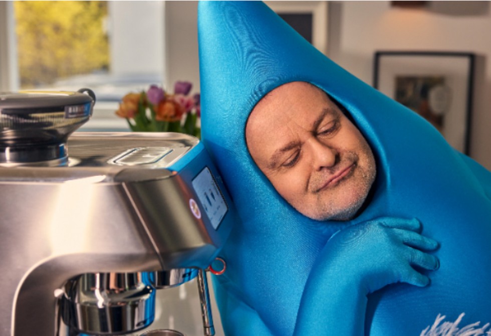 FLOWIAN, verkleidet als blauer Wassertropfen, an einer Kaffeemaschine in einer Küche
